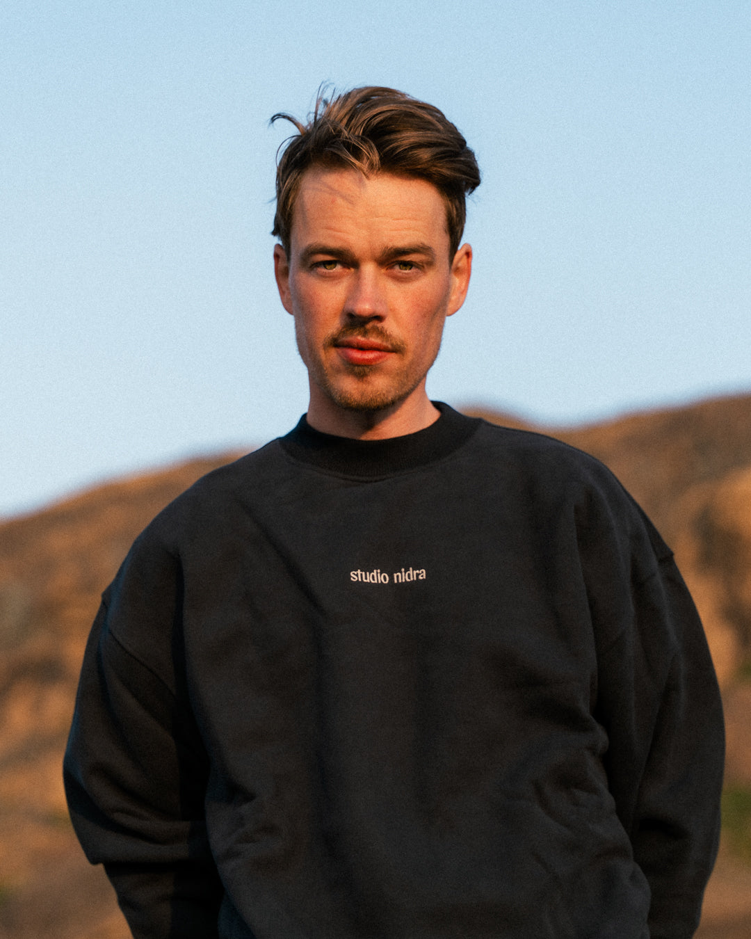 Man wearing The Crew Sweatshirt in black with Studio Nidra logo against mountainous background
